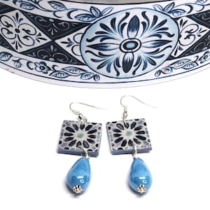 Può includere: Orecchini pendenti con parti superiori quadrate decorate e perle blu a goccia. Gli orecchini hanno ganci color argento e piccoli dettagli argentati. Il design sulla parte superiore quadrata è un motivo geometrico nei toni del blu e del grigio.