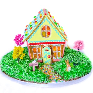 May include: A yellow gingerbread house decorated with colourful candies and frosting, surrounded by green grass and small trees. The house has a red door and windows, and a sign that says "Happy Easter".