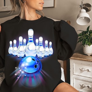 Puede incluir: Sudadera negra con un diseño de bolos dinámico. Una bola de bolos azul está frente a bolos blancos con detalles azules. El diseño incluye rayas de luz, creando una sensación dinámica y enérgica.