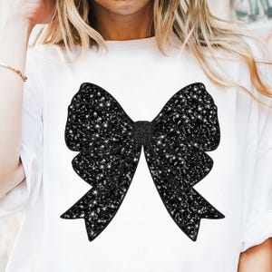Könnte beinhalten: Ein weißes T-Shirt mit einem schwarzen Glitzer-Schleifendesign.