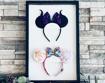 May include: Two pairs of character-themed Minnie Mouse ears displayed in a black frame. The top pair has black ears with purple glitter bows and horns. The bottom pair has white ears with a colorful floral design and gold accents.