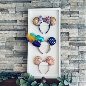 Può includere: Tre paia di orecchie di Minnie Mouse esposte su una cornice bianca. La coppia in alto ha un fiocco a motivi rosa e blu con orecchie di glitter dorate. La coppia in mezzo ha un fiocco viola con orecchie di glitter dorate e una fascia turchese. La coppia in basso ha un fiocco rosa con orecchie di glitter dorate.