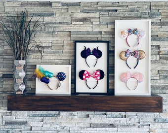 May include: Five pairs of Disney character ears displayed in white frames on a wooden mantel. The ears are decorated with various patterns and colors, including pink, purple, gold, and blue.