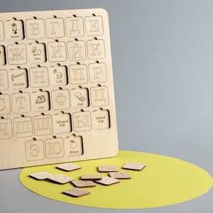 May include: A wooden alphabet puzzle with Russian letters and illustrations. The letters are arranged in a grid on a wooden board. The puzzle pieces are scattered on a yellow surface.
