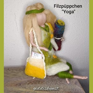 Könnte beinhalten: Eine Filzpuppe mit langen blonden Haaren, die ein grünes und gelbes Kleid trägt und eine kleine Tasche mit dem Text "Yoga Kids" trägt. Die Puppe hält ein zusammengerolltes Stück Stoff mit Regenbogenfarben. Die Puppe sitzt auf einer Holzoberfläche. Der Text "Filzpüppchen "Yoga"" ist über der Puppe geschrieben.