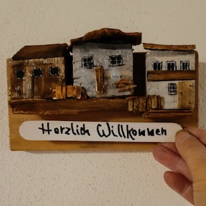 Könnte beinhalten: Ein hölzerner Wandbehang mit drei kleinen Häusern, die in Braun- und Grautönen bemalt sind. Die Häuser haben Fenster und Türen und sind in einer Reihe angeordnet. Das Schild unter den Häusern lautet "Herzlich Willkommen" in schwarzer Schrift.