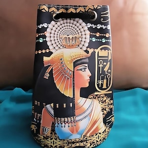May include: Black drawstring bag with a gold and blue print of an Egyptian queen wearing a crown and jewelry. The bag has a black drawstring and two gold eyelets.