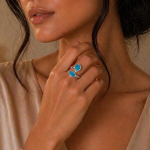 Può includere: Un anello d'argento con due pietre preziose blu brillante a forma di goccia. L'anello è indossato su una mano, con le pietre posizionate una accanto all'altra. Il design dell'anello è semplice, evidenziando il colore vibrante delle pietre.