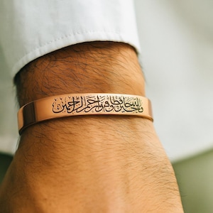 Può includere: Un bracciale a polsino in oro rosa con scritta araba incisa. Il bracciale è indossato su un polso, con una manica di camicia bianca sullo sfondo. Il testo inciso è nero.