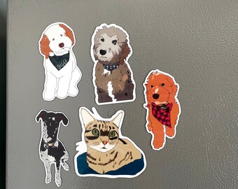 May include: Five refrigerator magnets featuring illustrations of dogs and a cat. The magnets are white with black outlines. The dogs are wearing bandanas. The cat is looking directly at the camera. The magnets are on a gray refrigerator.