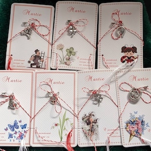Puede incluir: Ocho tarjetas blancas con un cordón rojo y blanco atado. Cada tarjeta tiene la palabra "Martie" impresa y un amuleto de plata diferente atado al cordón. Los amuletos incluyen un trébol, una herradura, una campana, un barrendero de chimeneas, una mariquita, una flor, una mariposa y un ramo de flores.