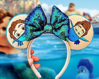 May include: A pair of light blue and tan mouse ears with a turquoise sequin bow. The ears feature embroidered images of a character from the animated film Luca.