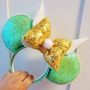 K&ouml;nnte beinhalten: Gr&uuml;ne und goldene Pailletten-Minnie-Maus-Ohren mit einer wei&szlig;en Schleife und einem wei&szlig;en Bommel oben.
