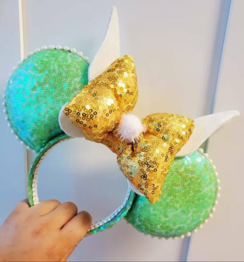 K&ouml;nnte beinhalten: Gr&uuml;ne und goldene Pailletten-Minnie-Maus-Ohren mit einer wei&szlig;en Schleife und einem wei&szlig;en Bommel oben.