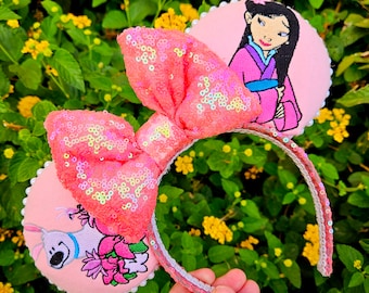 May include: Pink sequined headband with a large matching bow. The headband features two round ear designs, one with a cartoon image of Mulan and the other with a cartoon image of a dog and flowers. The headband is decorated with white pearls.