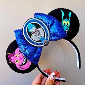 Puede incluir: Orejas de Minnie Mouse de Disney de color azul y negro con un lazo de lentejuelas azules y un parche bordado circular con Hades de la pel&iacute;cula de Disney H&eacute;rcules. La otra oreja presenta una serpiente marina bordada de color azul y verde.