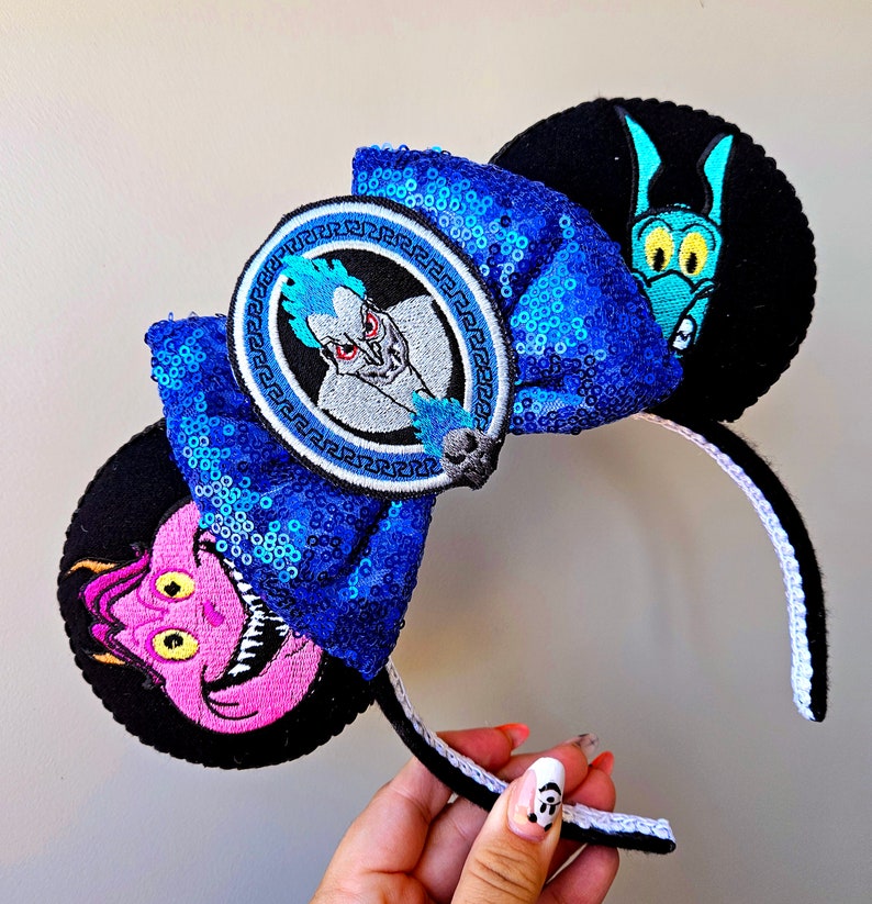 Puede incluir: Orejas de Minnie Mouse de Disney de color azul y negro con un lazo de lentejuelas azules y un parche bordado circular con Hades de la pel&iacute;cula de Disney H&eacute;rcules. La otra oreja presenta una serpiente marina bordada de color azul y verde.