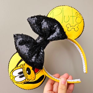 May include: Yellow felt Minnie Mouse ears with a black sequin bow and a yellow felt Pluto face with the text "Pluto" embroidered in green thread on the other ear.