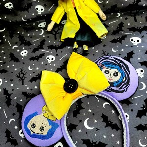 May include: Purple and yellow handmade mouse ears with a yellow bow and a black button. The ears feature an embroidered image of a girl with blue hair and a yellow dress.