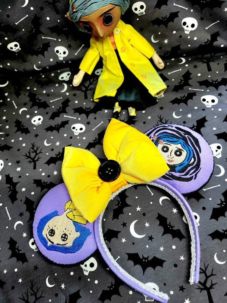 May include: Purple and yellow handmade mouse ears with a yellow bow and a black button. The ears feature an embroidered image of a girl with blue hair and a yellow dress.