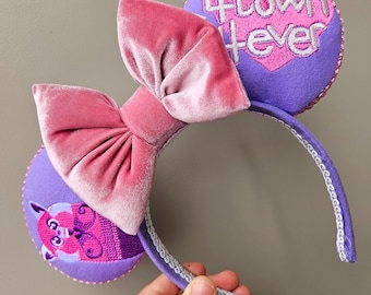 May include: Purple headband with a pink velvet bow and two purple ears. One ear has a pink heart with the text "Town Forever" embroidered on it. The other ear has a purple and pink cartoon character embroidered on it.