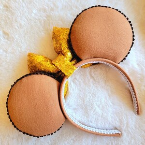 May include: Brown felt Minnie Mouse ears with a gold sequin bow. The headband is white with brown trim.