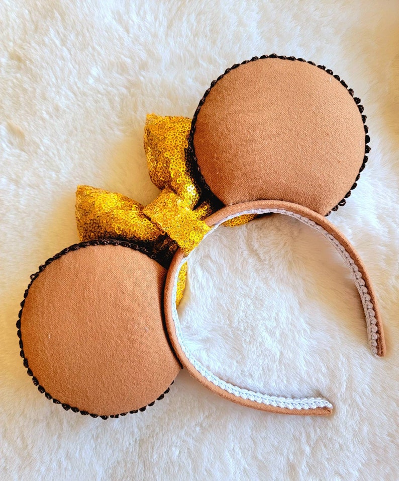 May include: Brown felt Minnie Mouse ears with a gold sequin bow. The headband is white with brown trim.
