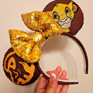 May include: A Simba-themed headband with a gold sequined bow and embroidered Simba faces on the ears. The ears are brown with a dark brown border. The headband is brown with a silver trim.