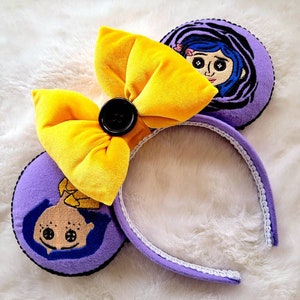 May include: Purple and yellow headband with a large yellow bow and a black button. The headband has two purple ears with embroidered images of a girl with blue hair and large eyes.