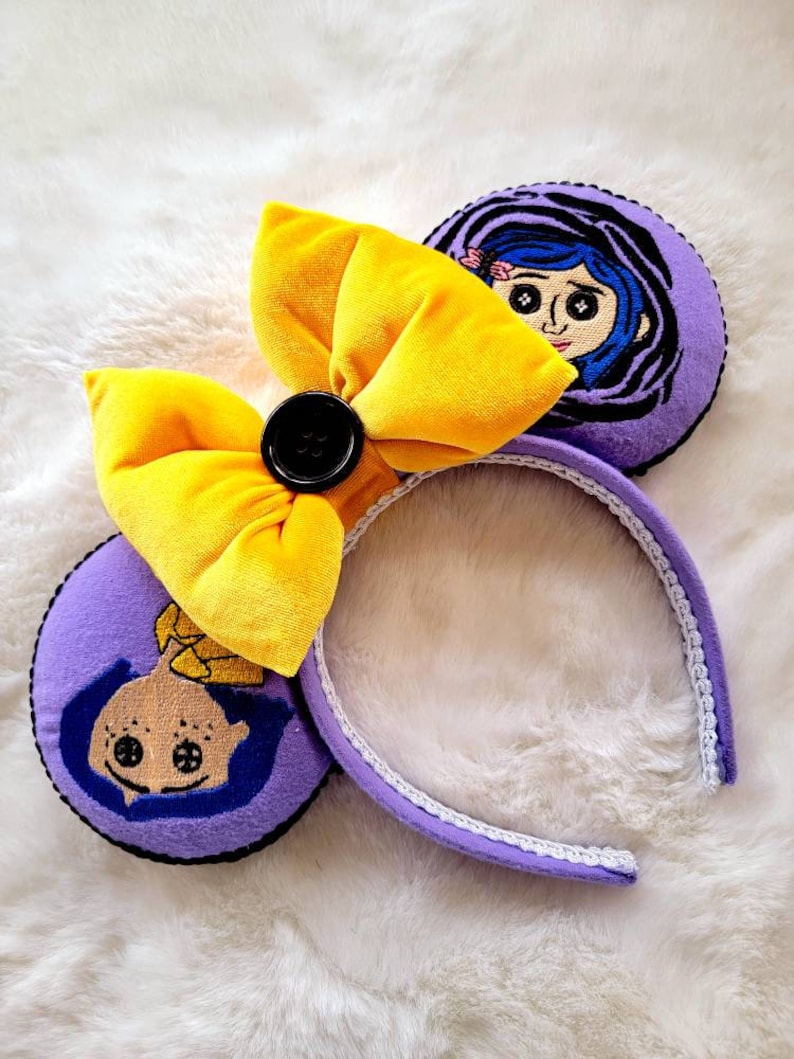 May include: Purple and yellow headband with a large yellow bow and a black button. The headband has two purple ears with embroidered images of a girl with blue hair and large eyes.