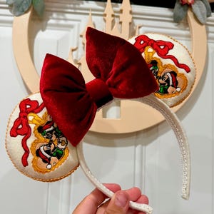 May include: Cream-colored mouse ears headband with embroidered cartoon characters in a gold frame. A large, plush, red velvet bow sits atop the headband. The ears have a gold trim and a red bow design. The headband is held by a hand.