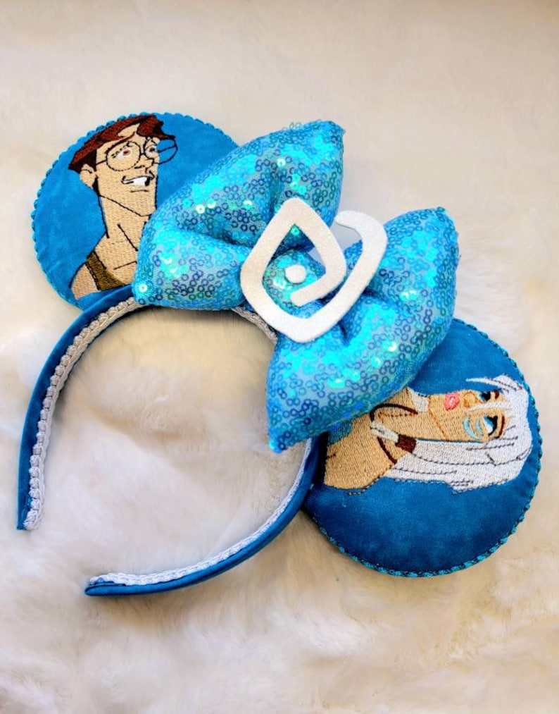 May include: A pair of blue and white mouse ears with a blue sequin bow and a white swirl design. The ears feature embroidered images of a man and a woman.