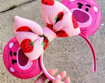 May include: Pink Minnie Mouse ears with a pink bow featuring a strawberry print and pink bear ears with embroidered faces.