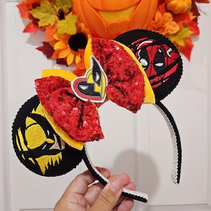 May include: Black and yellow mouse ears with a red and yellow bow. One ear features a black and yellow Wolverine character and the other ear features a black and red Deadpool character.