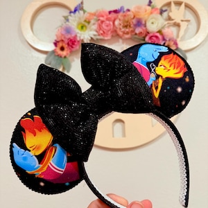 May include: Black Minnie Mouse ears with a sparkly black bow. The ears feature a colorful illustration of two characters from Disney's Elemental movie, a blue water character and a fiery orange character.