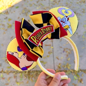 May include: Yellow headband with Roger Rabbit-themed ears. The ears feature cartoon graphics of Roger Rabbit and his love interest, Jessica Rabbit. A red, black, and yellow bow with the text "Roger Rabbit" is centered on the headband. The headband is held by a hand.