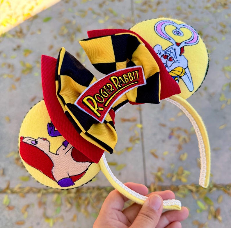 May include: Yellow headband with Roger Rabbit-themed ears. The ears feature cartoon graphics of Roger Rabbit and his love interest, Jessica Rabbit. A red, black, and yellow bow with the text "Roger Rabbit" is centered on the headband. The headband is held by a hand.