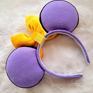 May include: Purple mouse ears with a yellow velvet bow. The headband is white with a black trim.