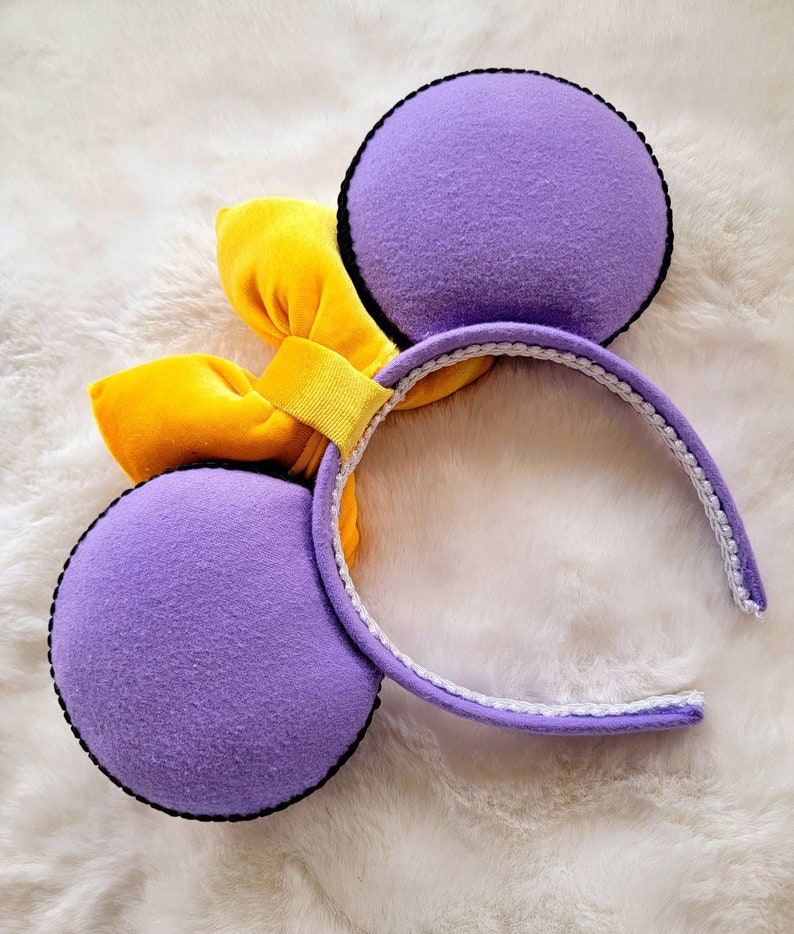 May include: Purple mouse ears with a yellow velvet bow. The headband is white with a black trim.