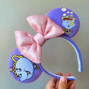 May include: Purple Minnie Mouse ears with a pink bow and embroidered images of the character Chip from Beauty and the Beast. The ears are decorated with white stitching and the character Chip is holding a teacup.