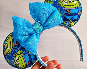 Puede incluir: Orejas de personaje azul y verde con un lazo azul de lentejuelas. Las orejas presentan un diseño de temática espacial con un personaje extraterrestre verde.