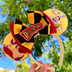 May include: Yellow mouse ears headband featuring Roger Rabbit characters. The headband has a black and yellow checkered bow with a "Roger Rabbit" patch. The ears have cartoon images of Roger Rabbit and Jessica Rabbit.