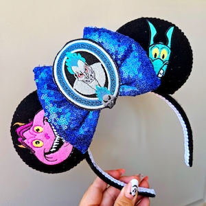 Puede incluir: Orejas de Minnie Mouse de Disney de color azul y negro con un lazo de lentejuelas azules y un parche circular con Hades de la pel&iacute;cula H&eacute;rcules. La otra oreja presenta un personaje de dibujos animados rosa y amarillo con cuernos.
