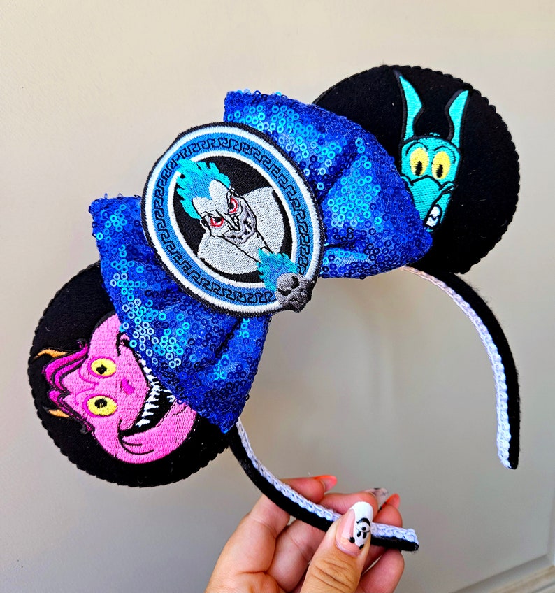 Puede incluir: Orejas de Minnie Mouse de Disney de color azul y negro con un lazo de lentejuelas azules y un parche circular con Hades de la pel&iacute;cula H&eacute;rcules. La otra oreja presenta un personaje de dibujos animados rosa y amarillo con cuernos.
