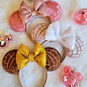 May include: Three pairs of Minnie Mouse ears with a concha design. The ears are brown, pink, and white with sparkly bows.