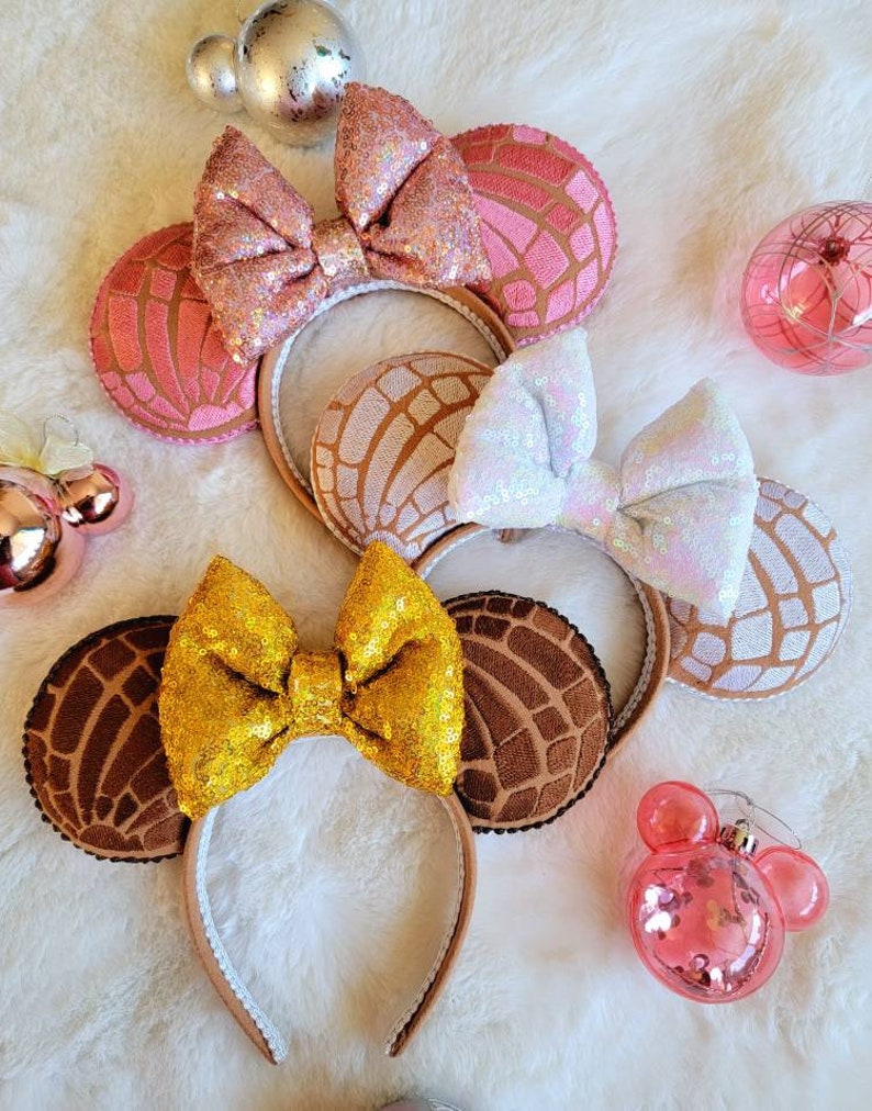 May include: Three pairs of Minnie Mouse ears with a concha design. The ears are brown, pink, and white with sparkly bows.
