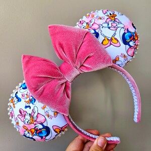 May include: Pink velvet bow headband with white Minnie Mouse ears featuring a print of Daisy Duck and Donald Duck. The ears are decorated with white pearls.