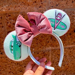 May include: A light blue headband with embroidered Cars-themed designs. The headband features a large, dusty rose velvet bow. One ear has a green car design, and the other has a "V8 Cafe" logo. The headband is held by a hand.