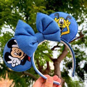 May include: Blue denim mouse ears headband with a large bow. One ear features Mickey Mouse, the other Pluto. The headband is white with blue trim. A fun accessory for theme park visits or cosplay.