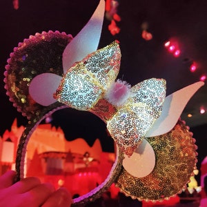 K&ouml;nnte beinhalten: Goldene und wei&szlig;e Pailletten-Minnie-Maus-Ohren mit einer gro&szlig;en wei&szlig;en Schleife und wei&szlig;en Hasenohren. Die Ohren sind mit kleinen, glitzernden Pailletten verziert.